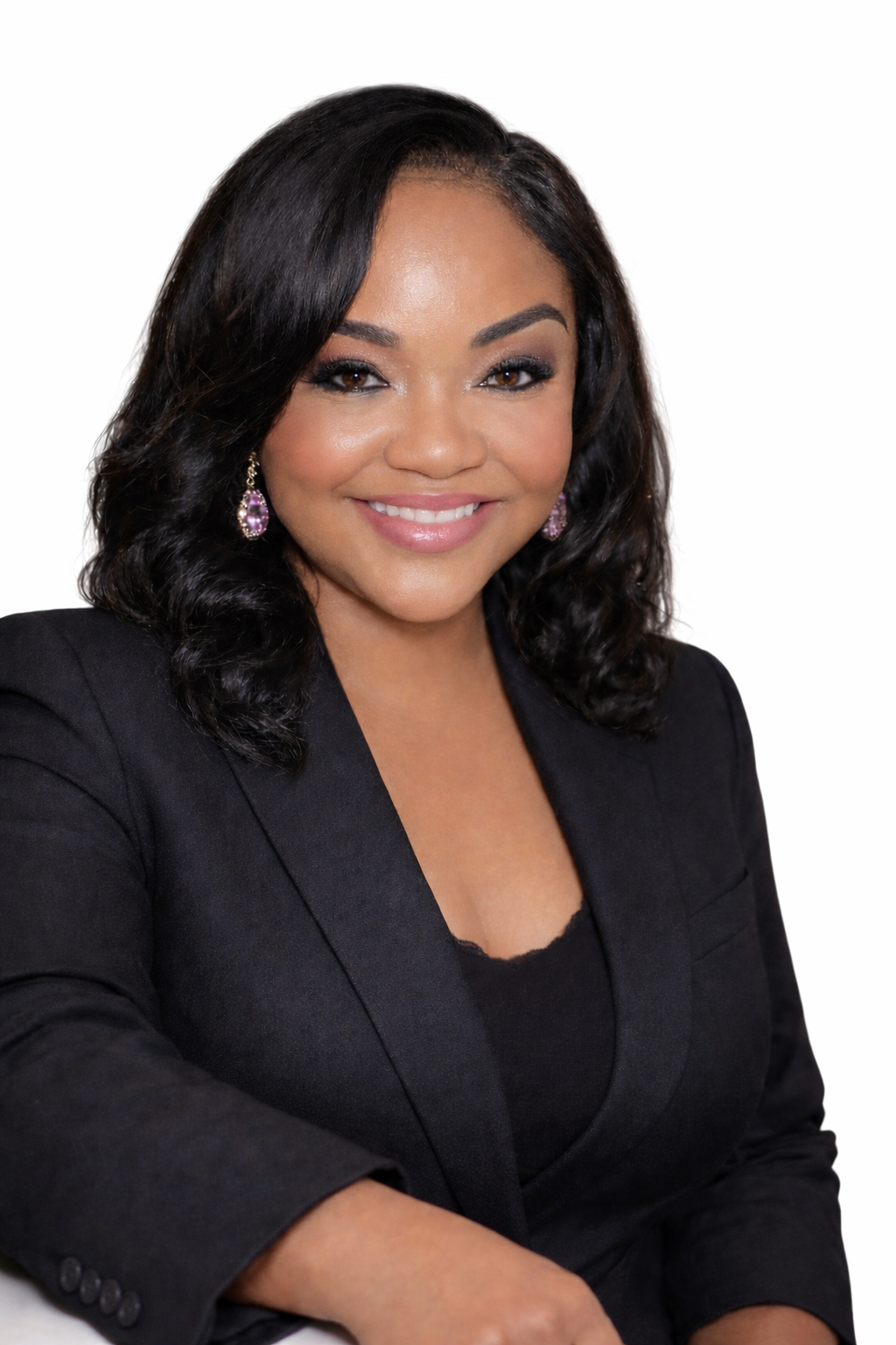 A woman with shoulder-length black hair, wearing a black blazer and purple earrings, smiling against a white background.