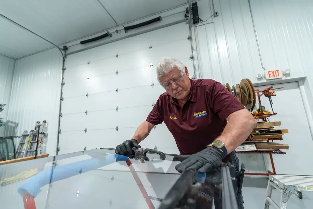 Kevin working on a glass panel in a garage or workshop. He is wearing black gloves and a maroon shirt with Southeastern Glass Co. Logo, with various tools and storage shelves visible in the background.