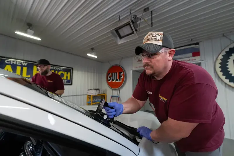 Two men working on a car inside a garage or workshop. The man in the foreground is wearing a cap, glasses, blue gloves, and a maroon shirt, and is replacing the car's windshield with a tool.