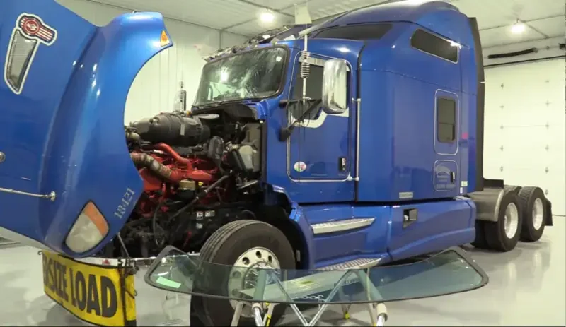 Blue semi-truck with hood open in a garage, displayed with a windshield in front of it.