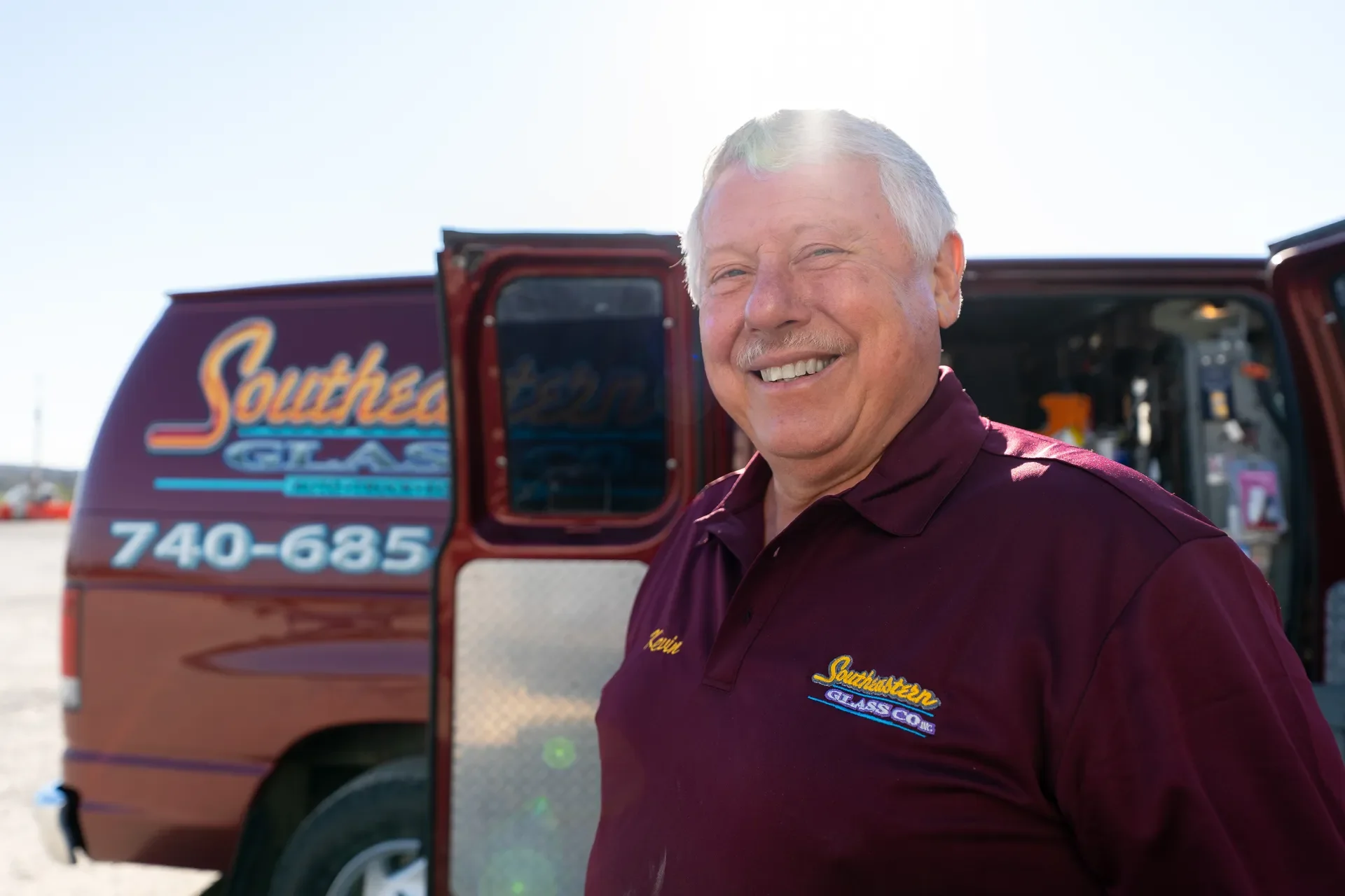 Kevin smiling wearing a maroon shirt with a logo from Southern Glass, standing outdoors in front of a red service van with the same logo. The van has its back door open, revealing tools and equipment inside. The background is bright and sunny.