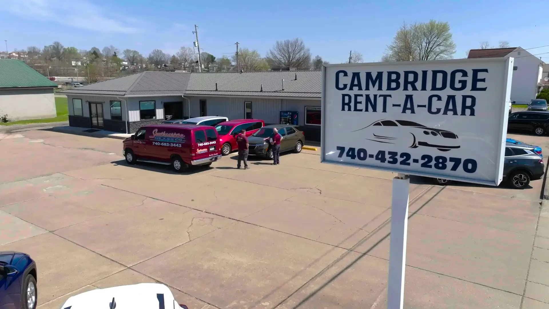 Parking lot outside Cambridge Rent-A-Car with a large sign, multiple cars, and two employees standing near a gray car.