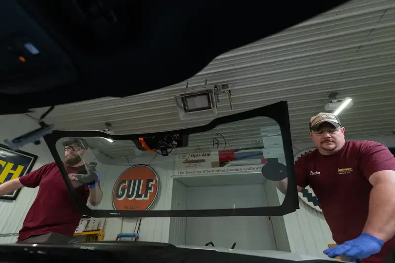 Two men with glasses, one wearing a cap, working inside a garage, replacing a windshield, shot from car interior.