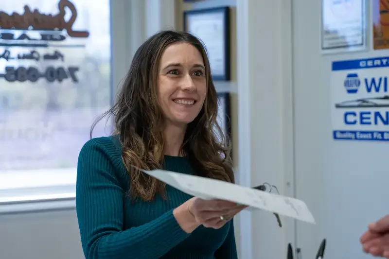 A woman with long brown hair smiling and handing a piece of paper to someone in an office setting.