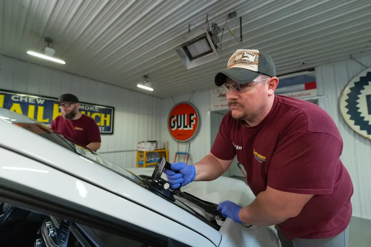 A man wearing a gray cap, safety glasses, and blue gloves inspects a car's windshield at Southeastern Glass Co.