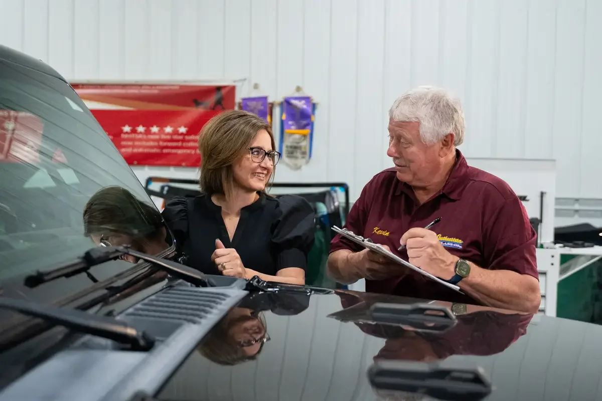 Candi and Kevin standing next to a black vehicle inside an Southeastern Glass Co glass repair shop, with the woman smiling and looking at the man, who is wearing a maroon shirt and holding a clipboard, engaging in conversation.