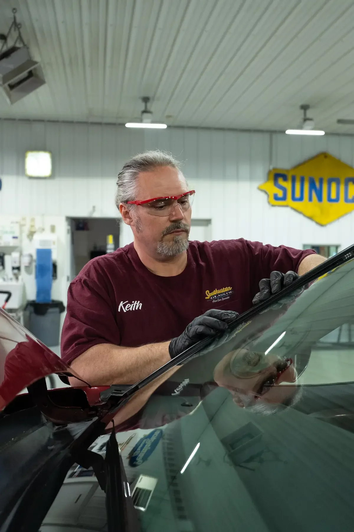 Keith wearing red glasses, black gloves, and a maroon shirt with the name 'Keith' is working on a car in an auto repair shop.