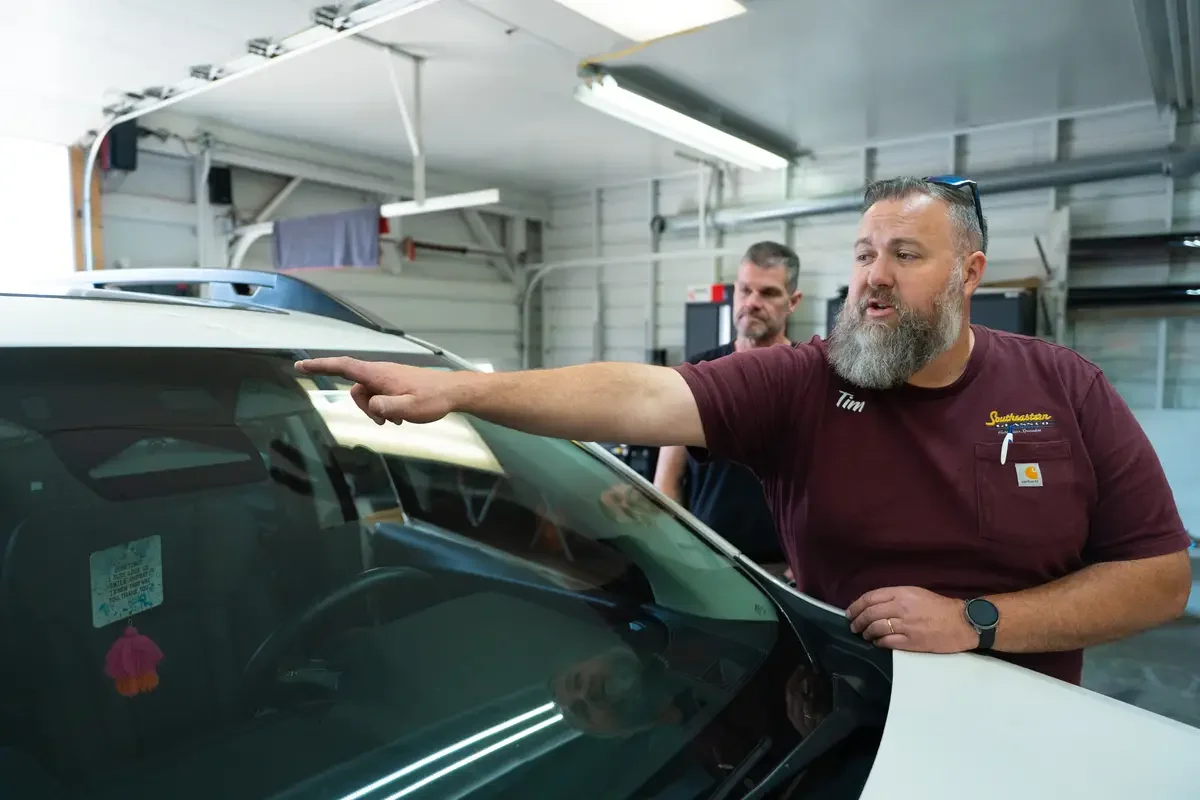 Tim, a glass expert with a beard and glasses on his head, points at a car's windshield while another man looks on inside a workshop or garage.
