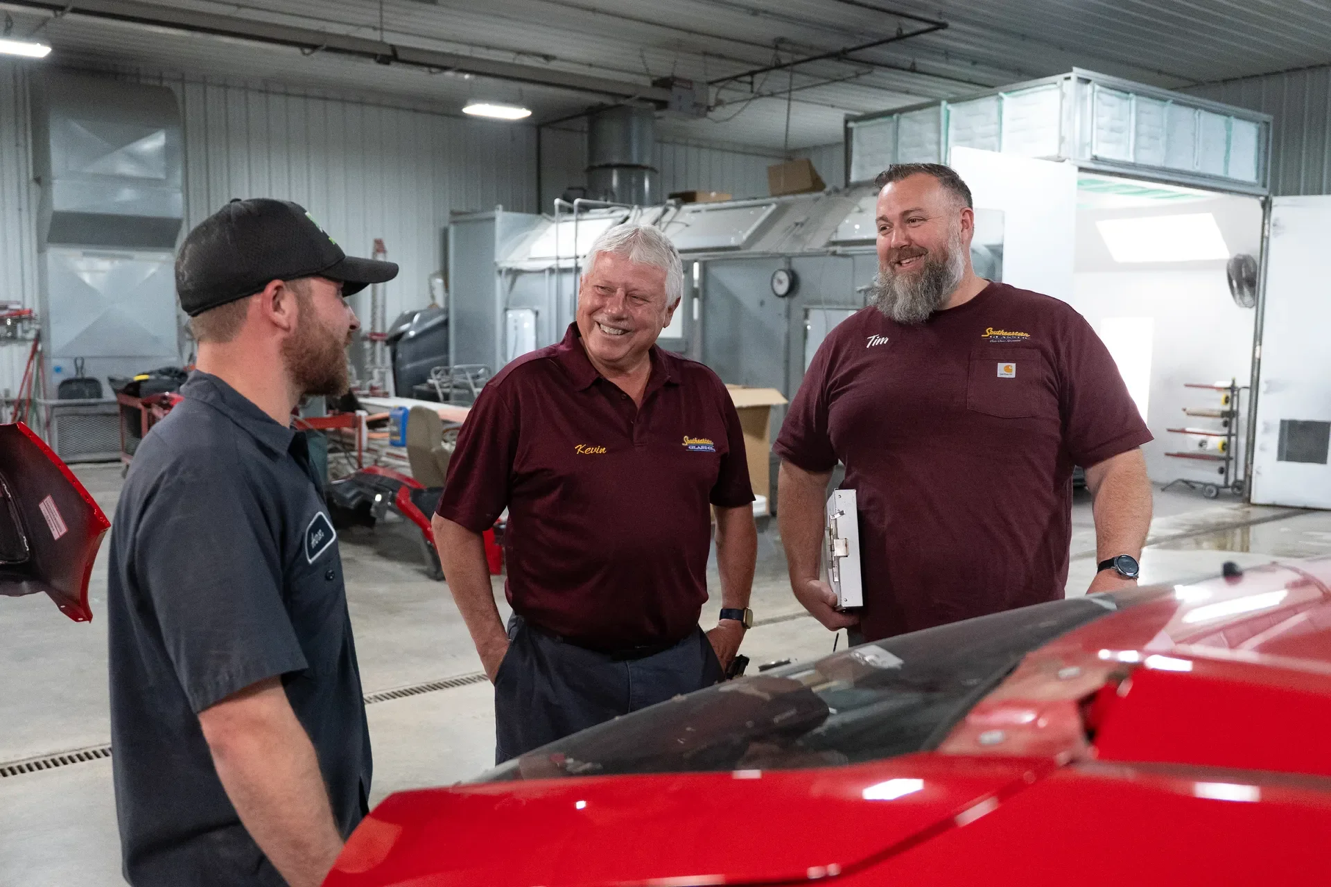 Three glass professionals are engaging in a conversation inside a workshop. They are standing near a red car, surrounded by workshop tools and equipment.