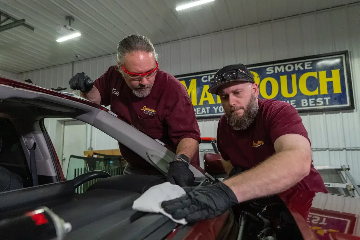 Two men working together on a car in a glass repair shop. One man is using a cloth to clean the car's dashboard while the other looks on. Both men wear maroon shirts and black gloves.