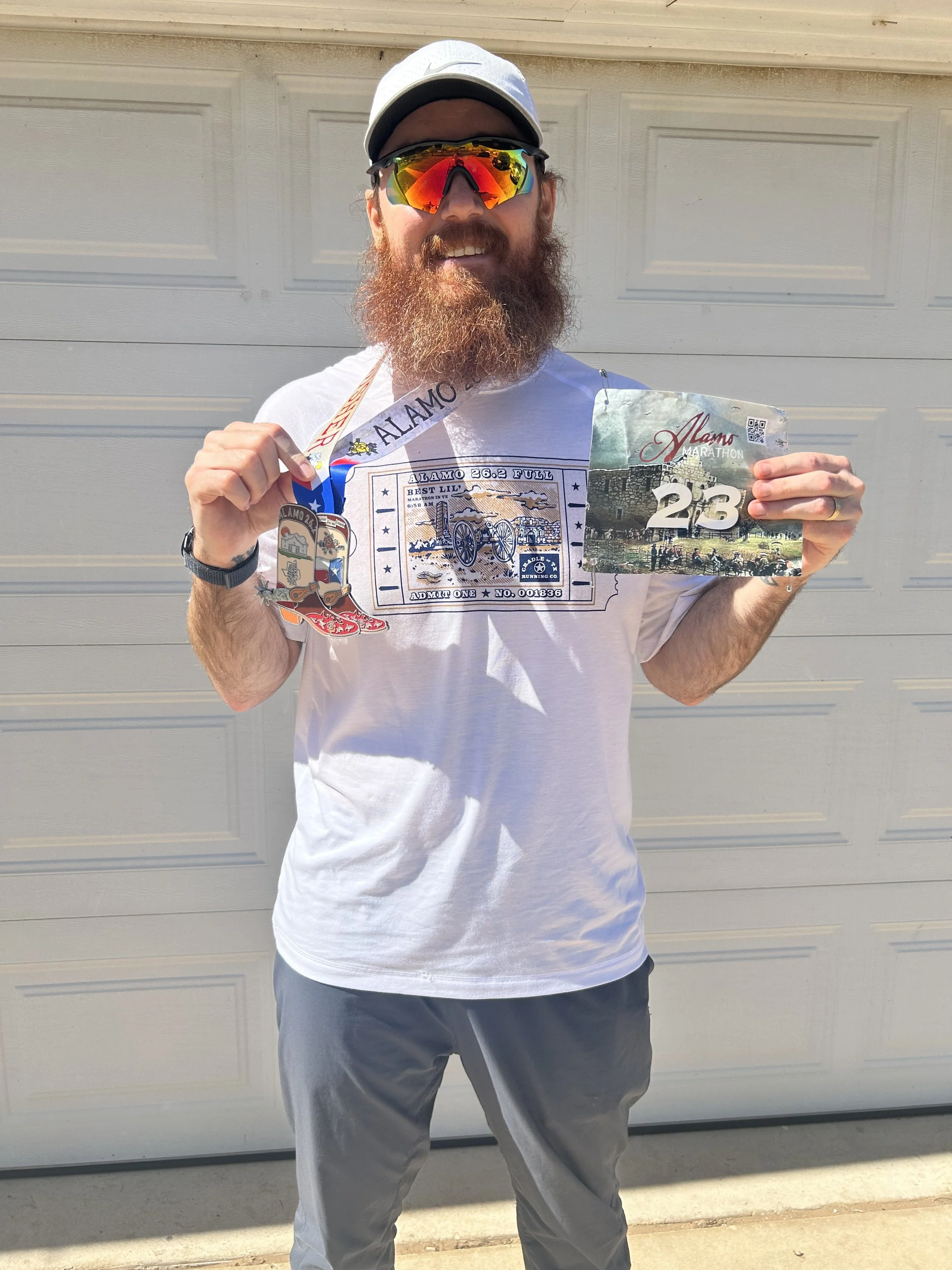 A man with a full beard is smiling and holding a marathon bib numbered 23, wearing sunglasses, a white cap, a white T-shirt, and gray shorts, standing in front of a gray garage door.