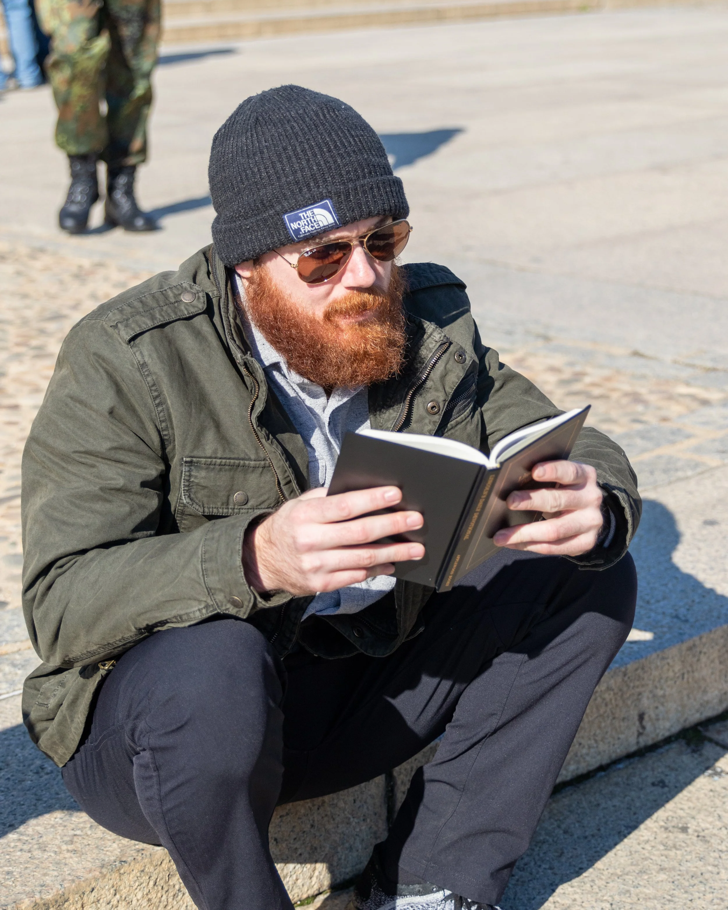 A man with a red beard wearing a black beanie, sunglasses, and an olive green jacket sitting outdoors on concrete steps, reading a book.