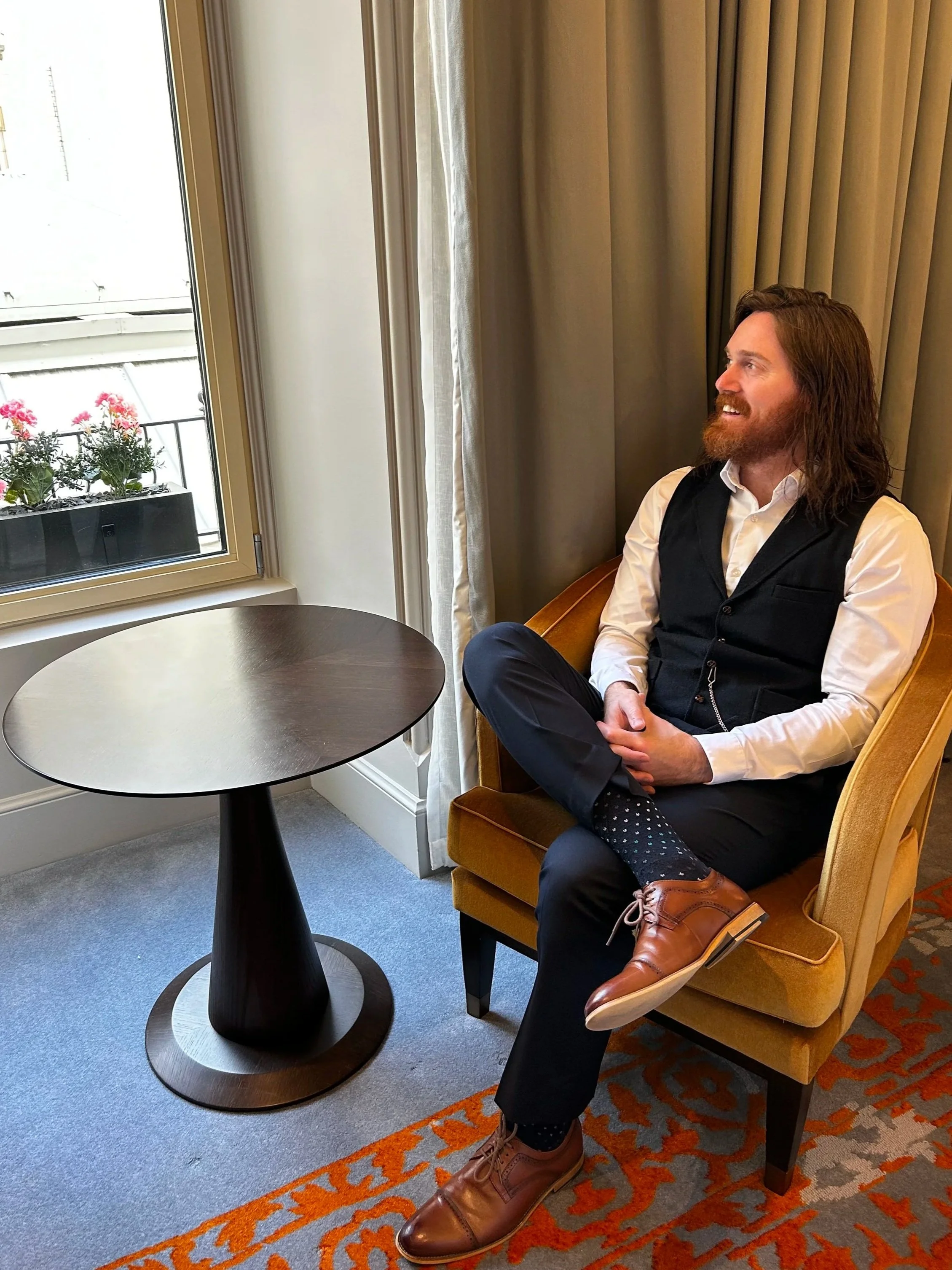 A man with long hair and a beard wearing a white shirt, black vest, and dark trousers sitting in a yellow armchair next to a window with beige curtains, with a small round table in front of him. He has brown shoes and patterned socks, and is looking toward the window with a smile.