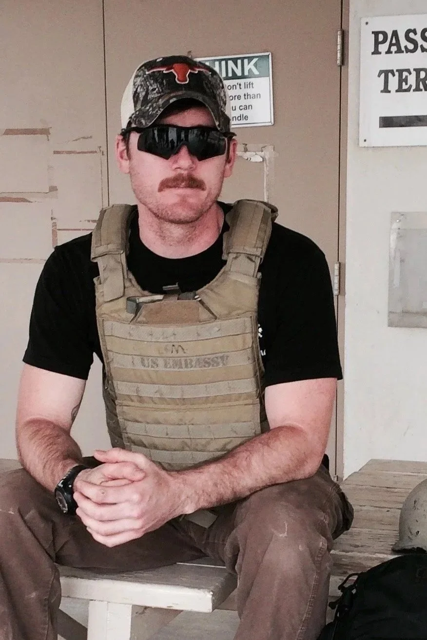 A man with a mustache sitting on a bench, wearing a camouflage hat with a Texas Longhorns logo, large black sunglasses, a tan tactical vest, a black T-shirt, brown pants, and a watch. He has his hands clasped together and is in an indoor setting with signs and a door in the background.