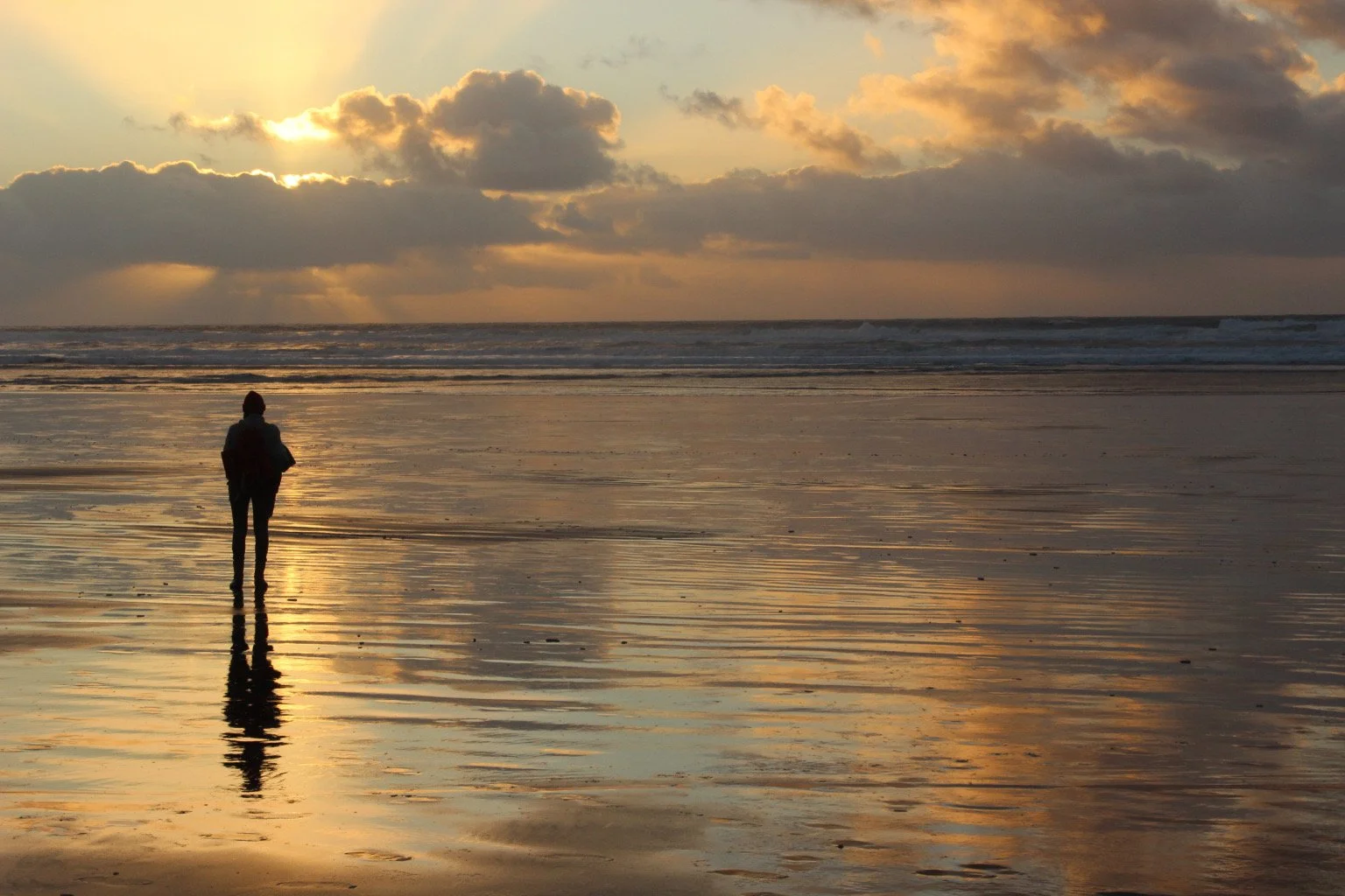 A person standing on wet sand at the beach during sunset, reflecting the sky with clouds and the sun's rays
