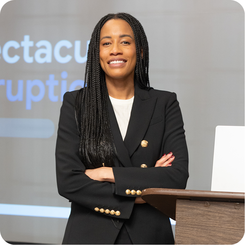 A woman with braids wearing a black blazer with gold buttons, standing confidently with arms crossed in front of a presentation screen.