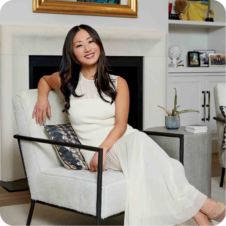 A woman with long dark hair sitting on a modern white armchair in a living room, smiling at the camera, with a fireplace, a painting above, books, and decorative items visible in the background.