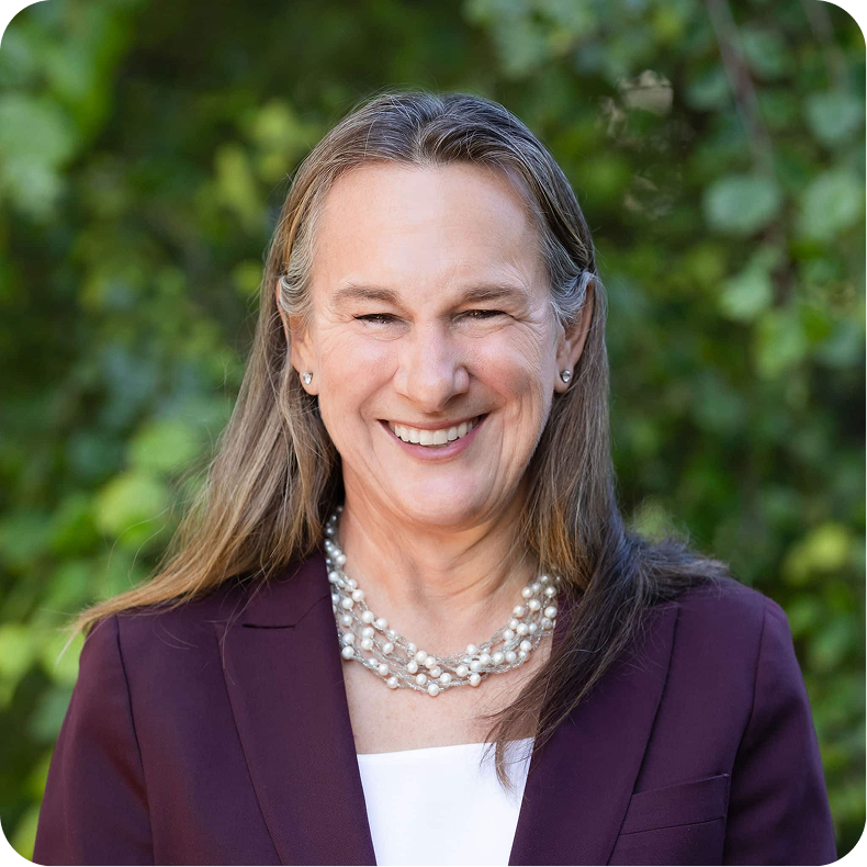 A middle-aged woman with long light brown hair, smiling in front of green foliage, wearing a dark purple blazer, white top, and multiple strands of pearl necklaces.