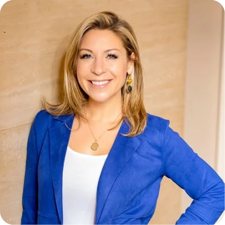A smiling woman with blonde hair wearing a blue blazer, white top, and gold earrings, standing against a beige wall.