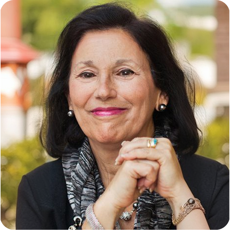 A smiling woman with black hair, wearing a black blazer with a patterned scarf, earrings, and a ring, sitting outdoors with her hands clasped.