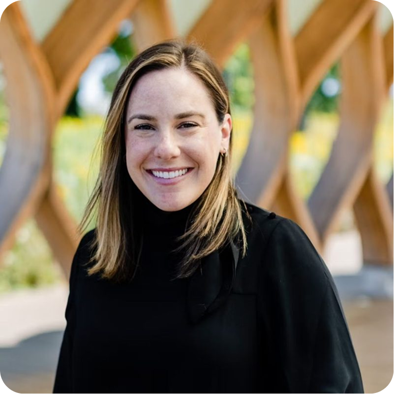 A smiling woman with shoulder-length light brown hair wearing a black top, outdoors with wooden arches in the background.