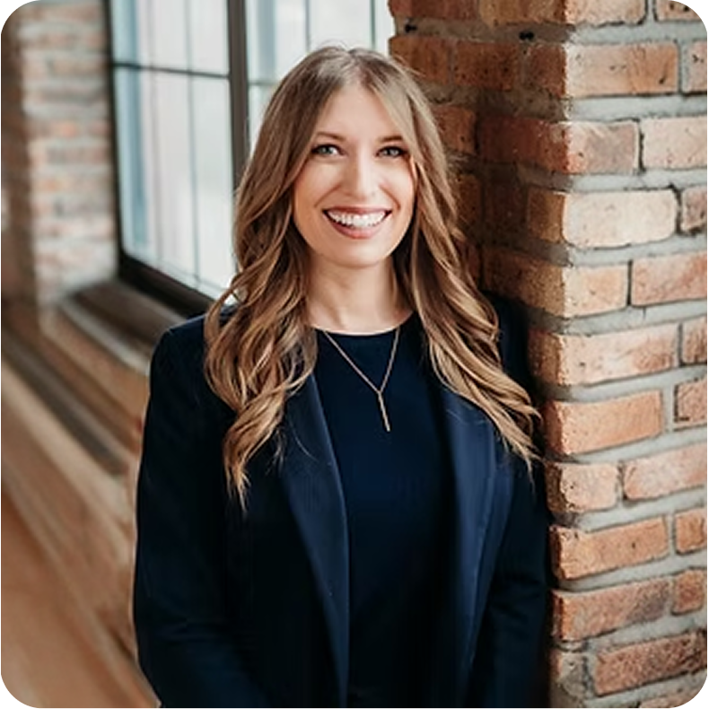 A woman with long wavy blonde hair, wearing a dark blazer and necklace, smiling and leaning against a brick wall near a window in a modern office or apartment.