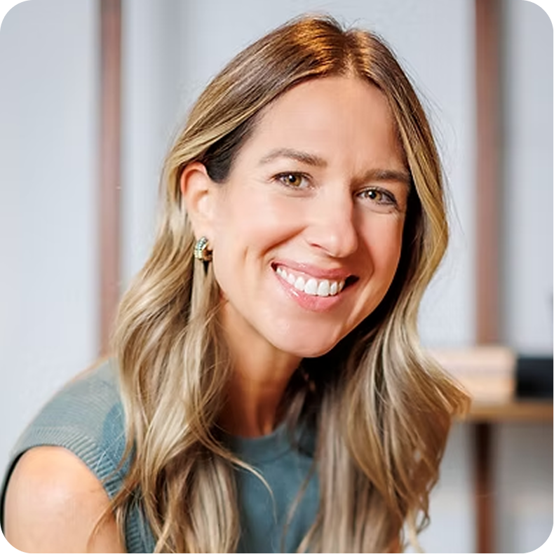 Portrait of a smiling woman with blonde hair wearing a teal sleeveless top, in a modern office setting.