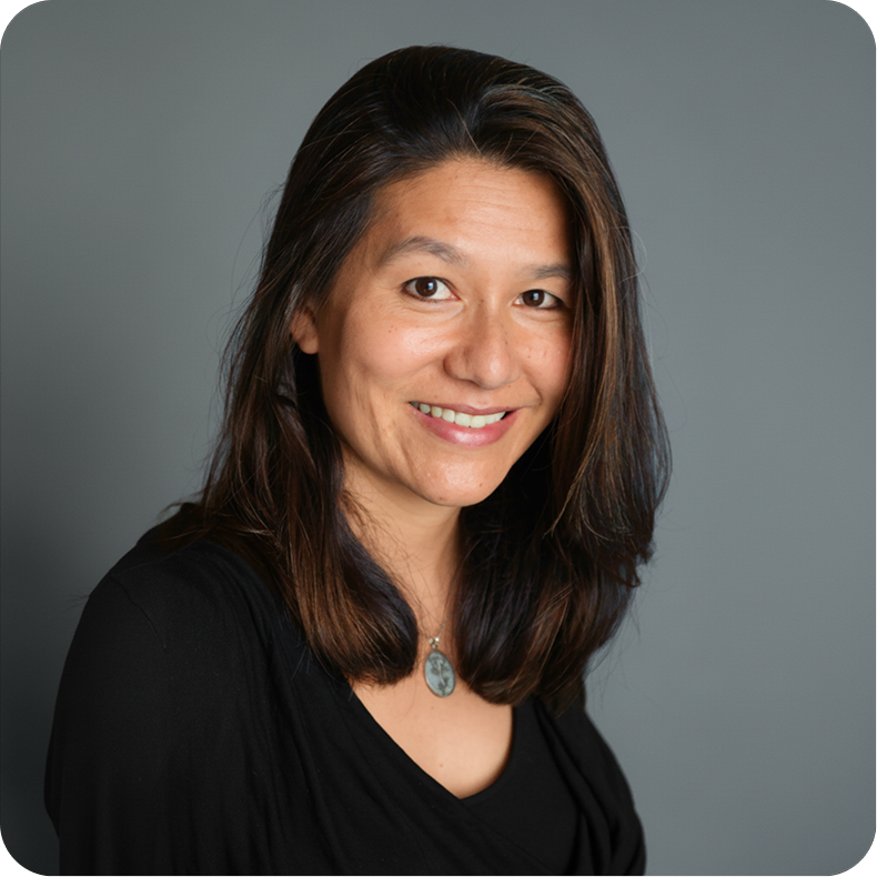 Portrait of a woman with shoulder-length dark brown hair, smiling, wearing a black top and pendant necklace, against a gray background.