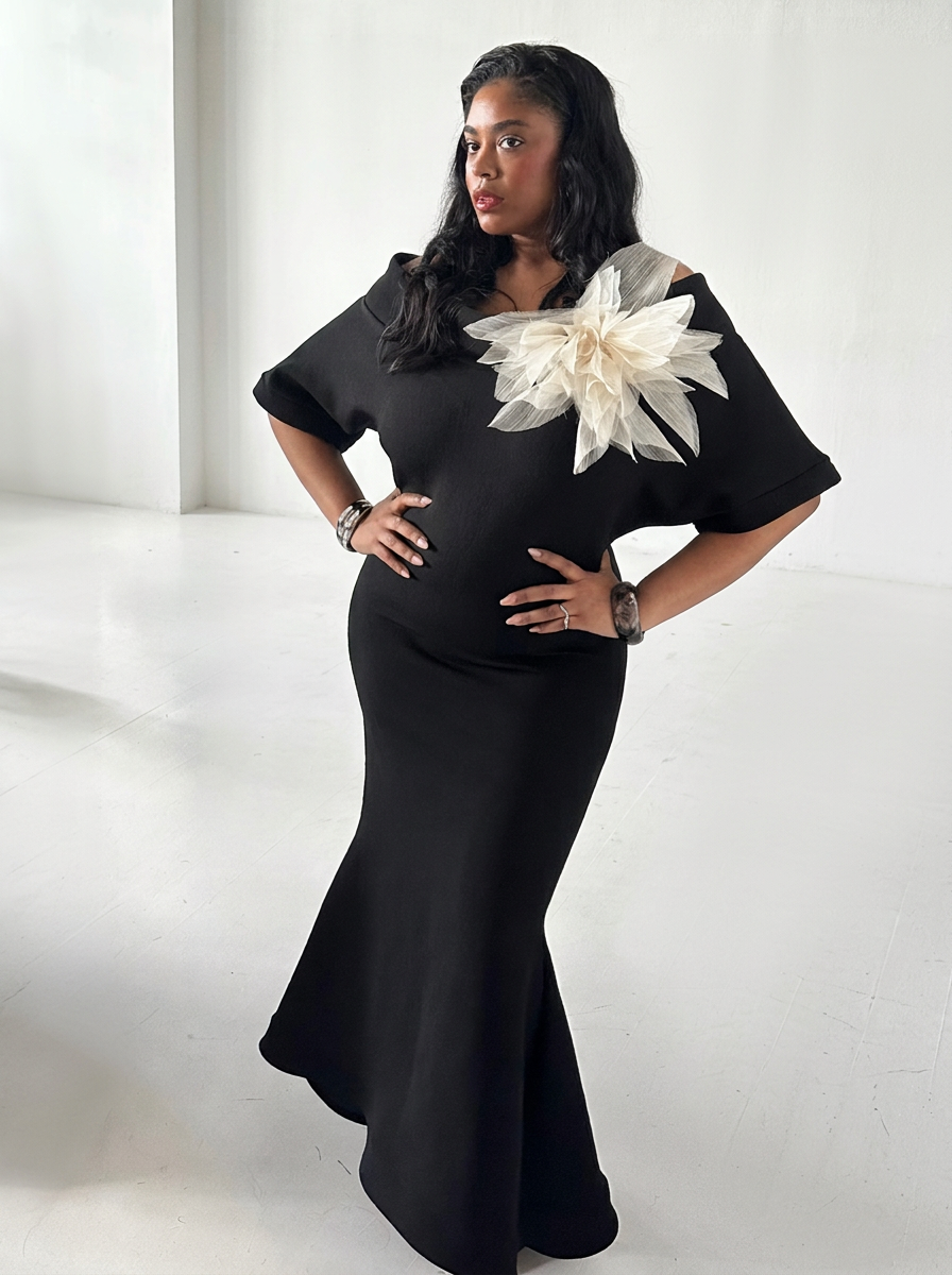 A sculptural elegance, the look pairs a sleek black silhouette with an extravagant ivory floral detail that commands attention across the shoulder. The softly draped neckline and voluminous petals create a striking contrast against the streamlined go