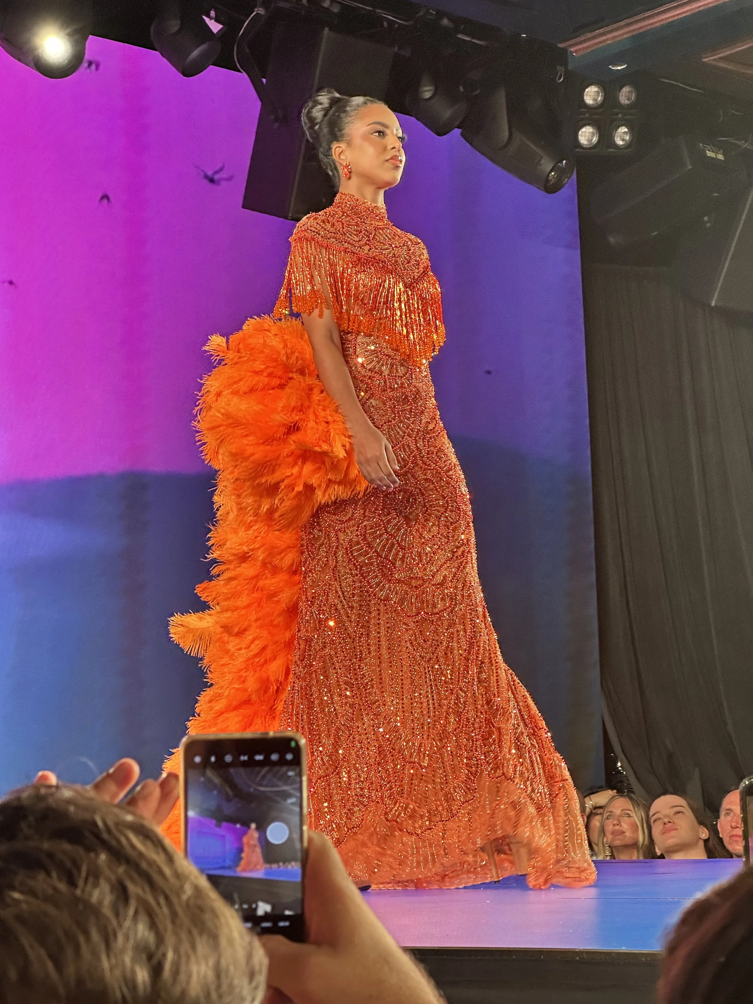 A richly beaded gown is paired with an enormous feathered train. The contrast between the structured, heavily embellished dress and the exuberant orange plume gives the look a strong performance quality. 
