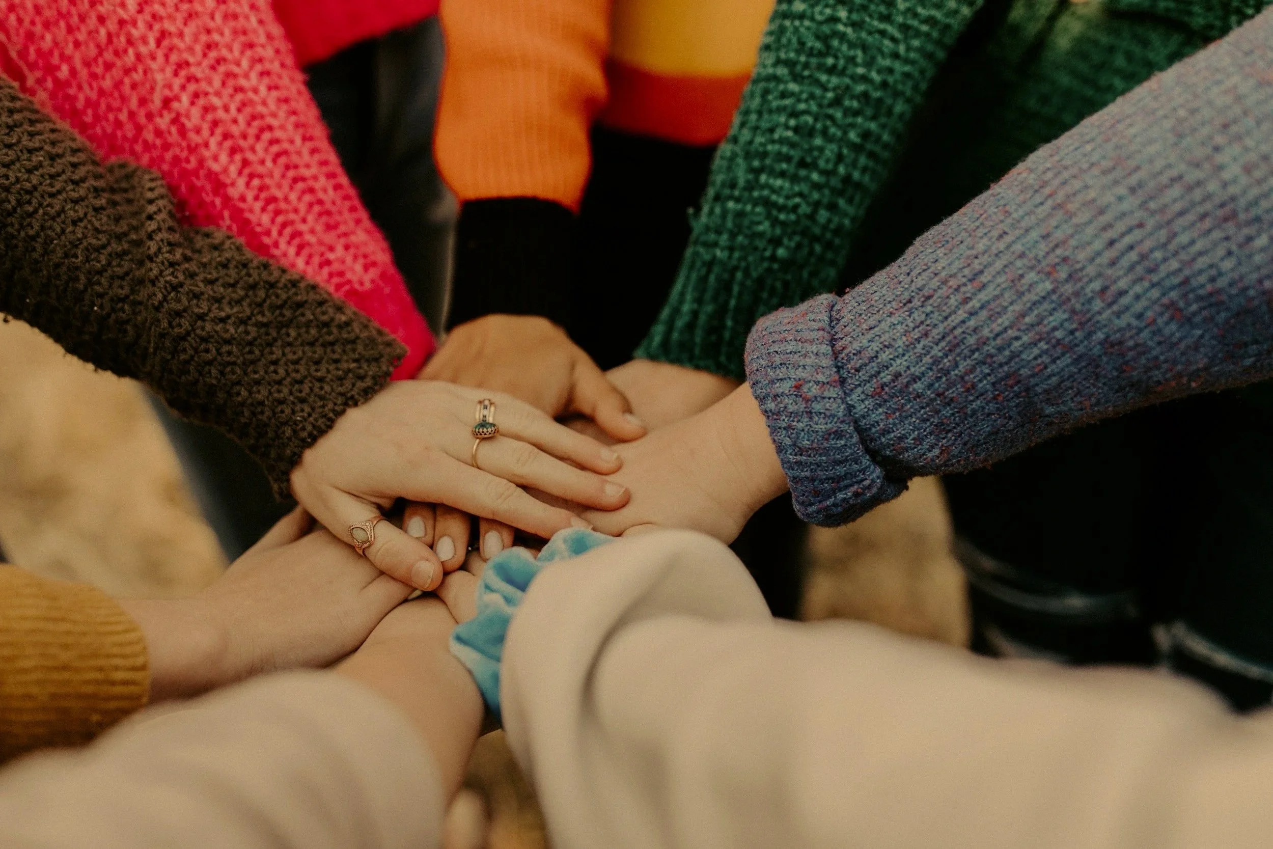 Multiple people holding hands in a circle, with focus on their hands and wrists, wearing colorful knitted sweaters.
