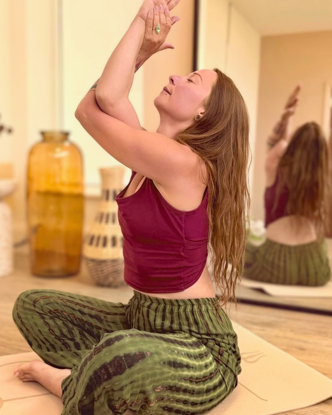 A woman practicing yoga indoors, sitting on a mat with her eyes closed and hands in a prayer position above her head, wearing a sleeveless top and loose pants.