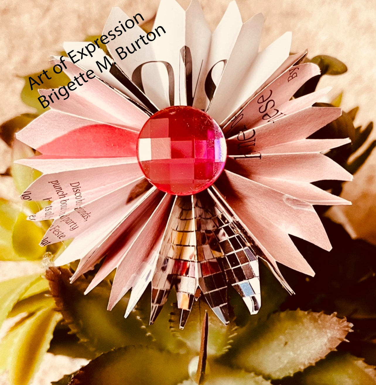 A handcrafted paper flower with pink and white petals, decorated with a pink, faceted, gem-like center and a reflective, mosaic-patterned metallic base.