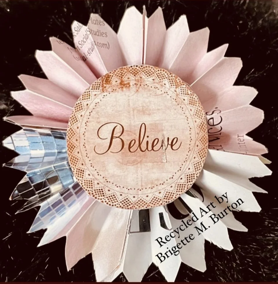 An art piece with a pink circular center that has the word "Believe" written on it, surrounded by layered paper petals with recycled materials, created by Brigette M. Burton.