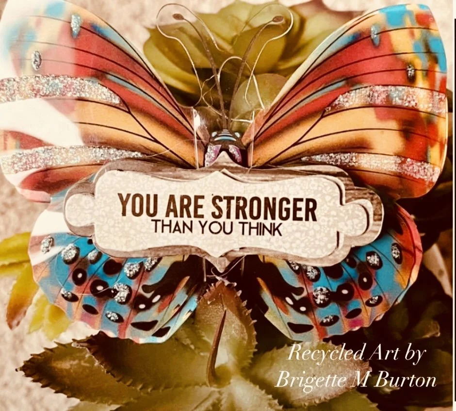 A decorative butterfly with colorful, glittery wings holding a sign that reads "You Are Stronger Than You Think." The butterfly is part of recycled art by Brigette M. Burton, featuring vibrant and shiny materials.