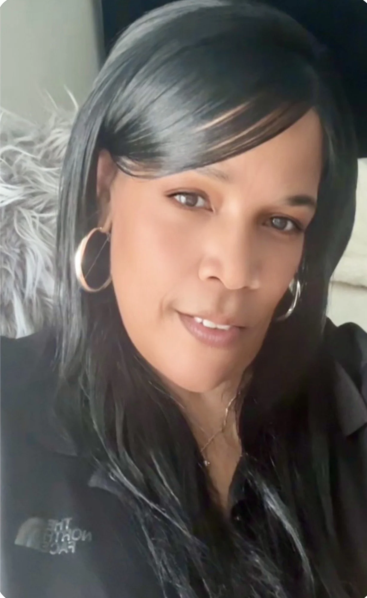 Close-up of a woman with long black hair, hoop earrings, and a black jacket, smiling slightly.