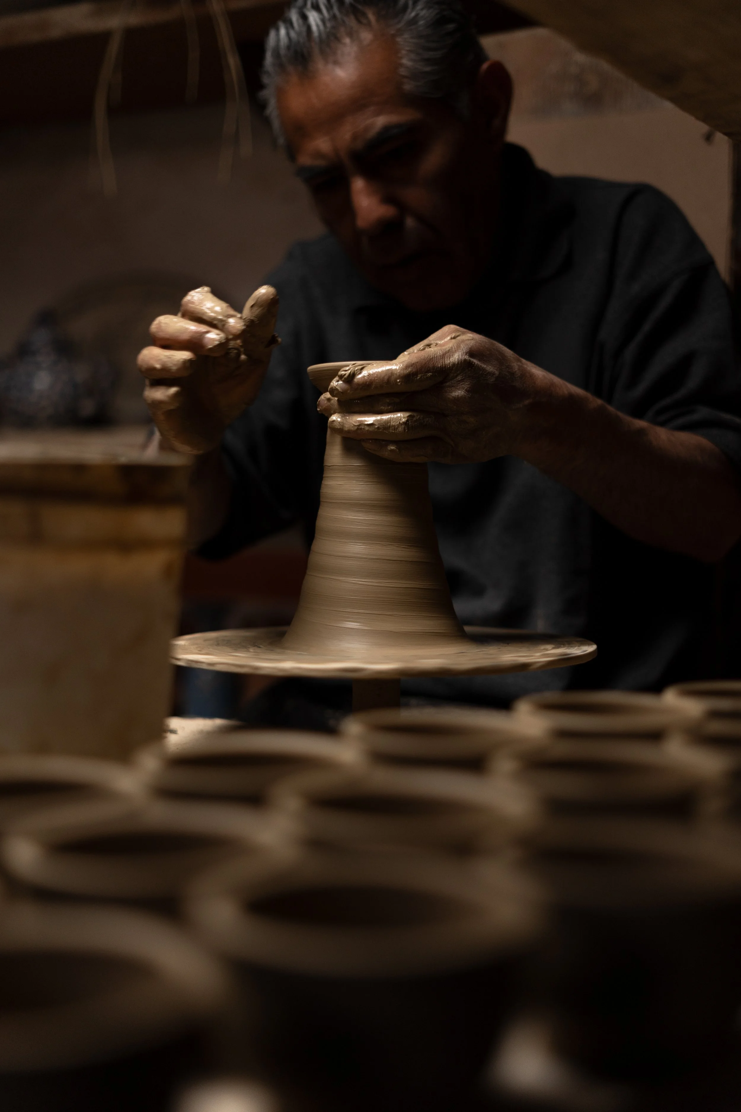 Un artesano tallando una vasija de cerámica en un taller, con múltiples vasijas en primer plano y fondo.