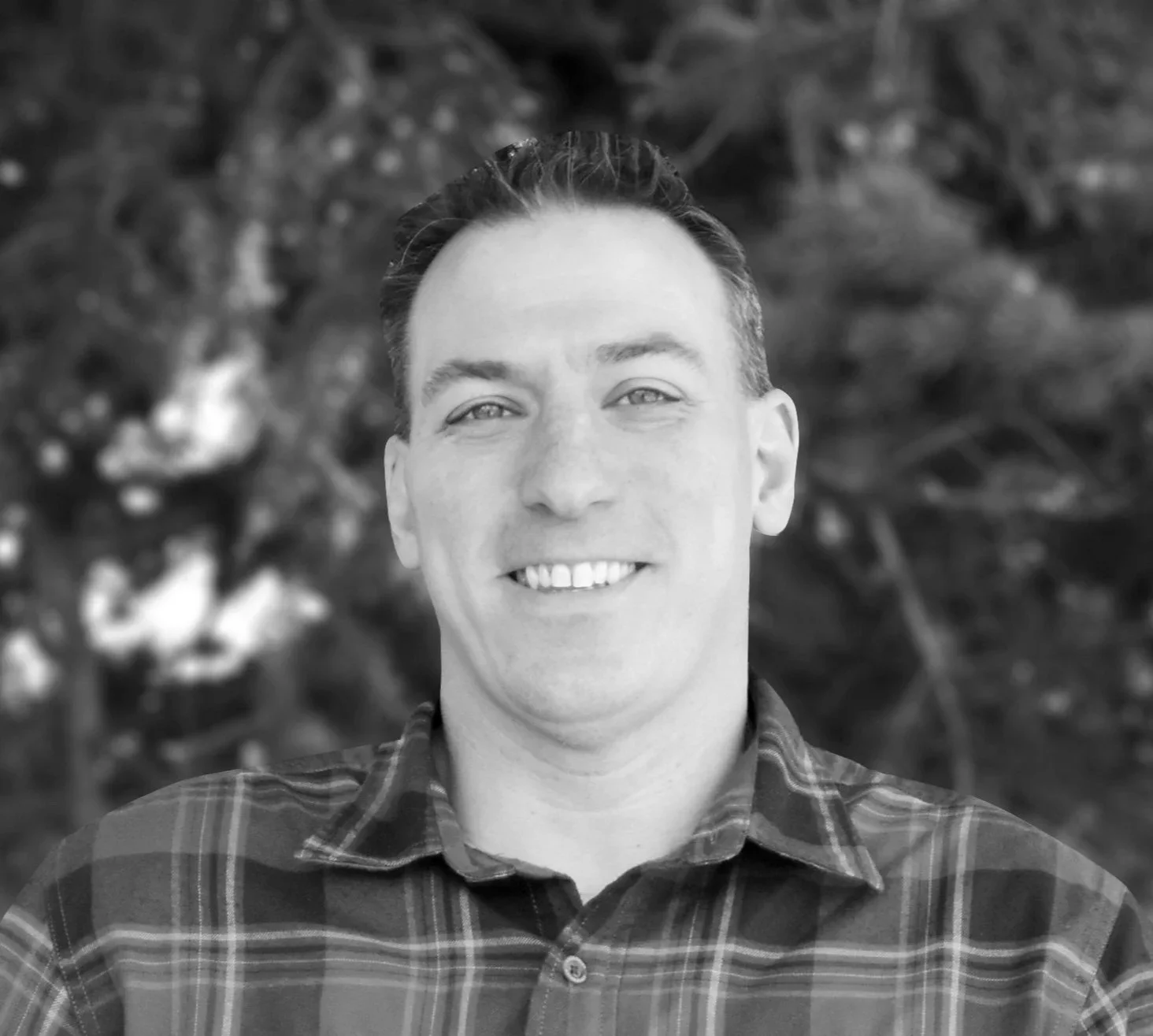 Black and white photo of a smiling man with short hair, wearing a checkered shirt, outdoors with blurred foliage in the background.