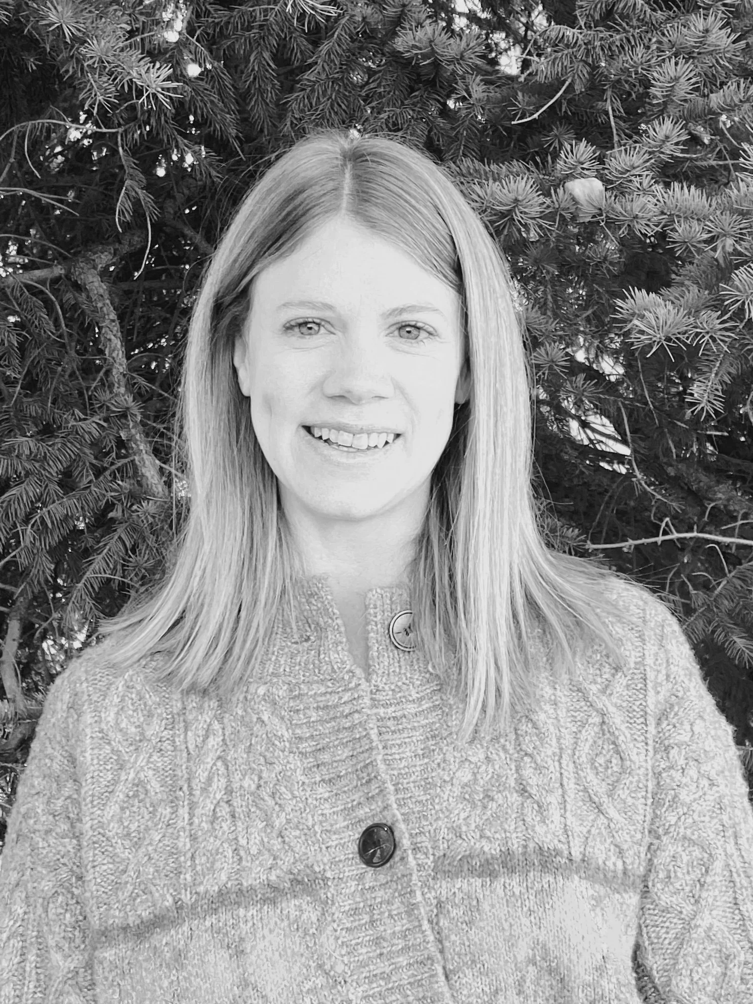 A woman with long blonde hair wearing a textured sweater and large buttons, standing outdoors in front of pine tree branches.