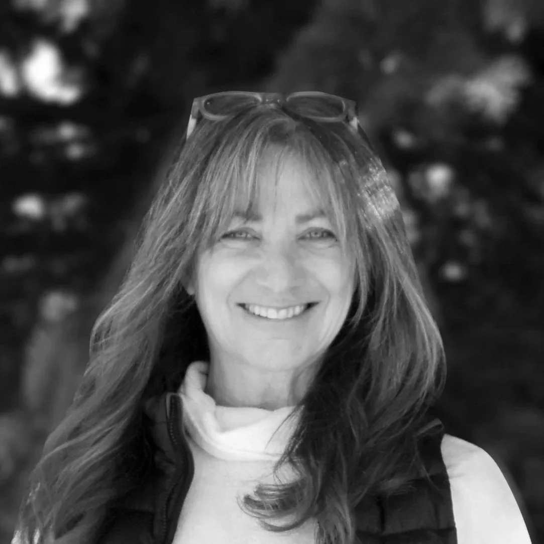 Black and white image of a woman with long hair smiling, wearing glasses on her head and a vest.