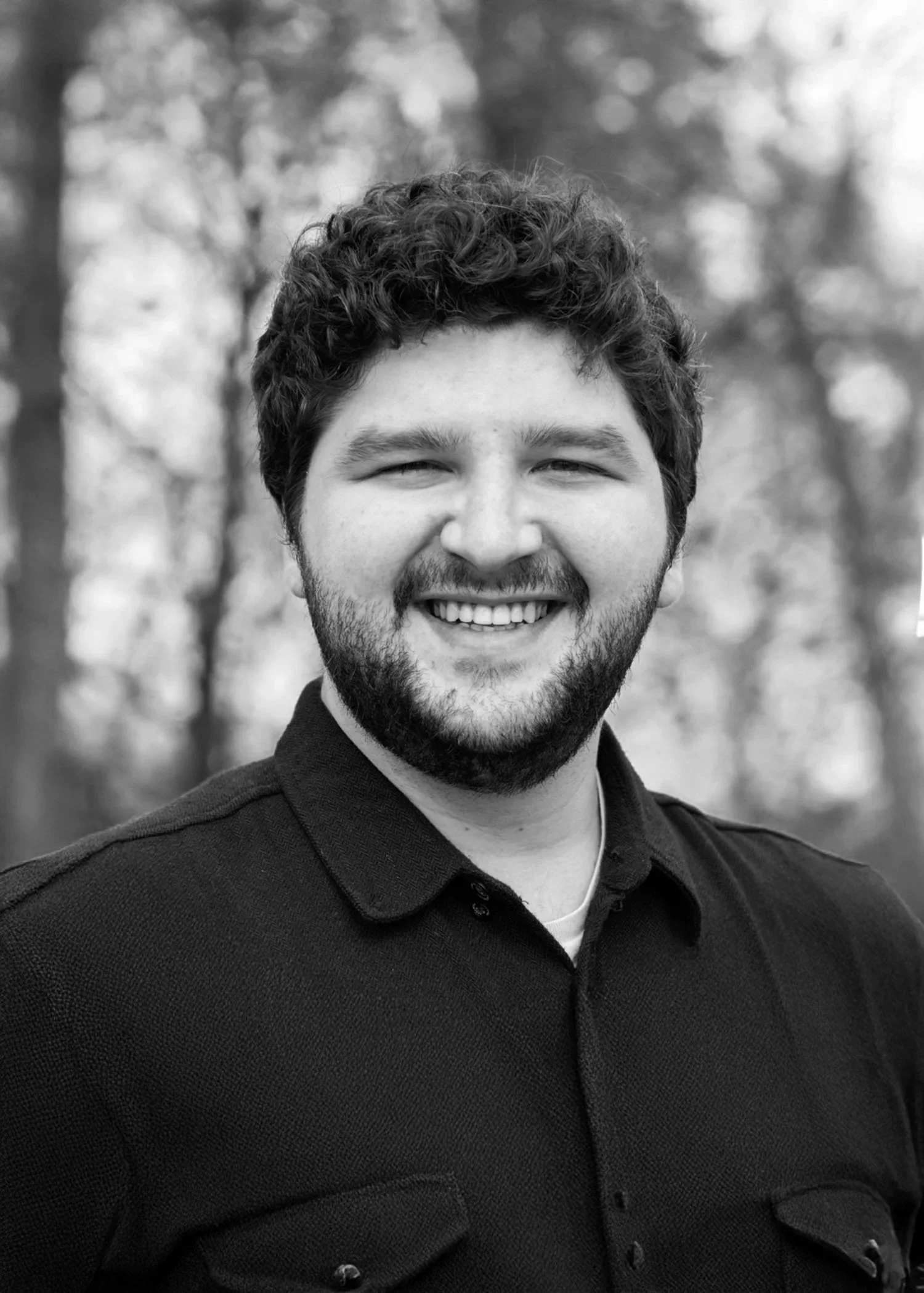 Black and white portrait of a smiling man with curly hair and a beard, wearing a dark jacket, outdoors with blurred trees in the background.