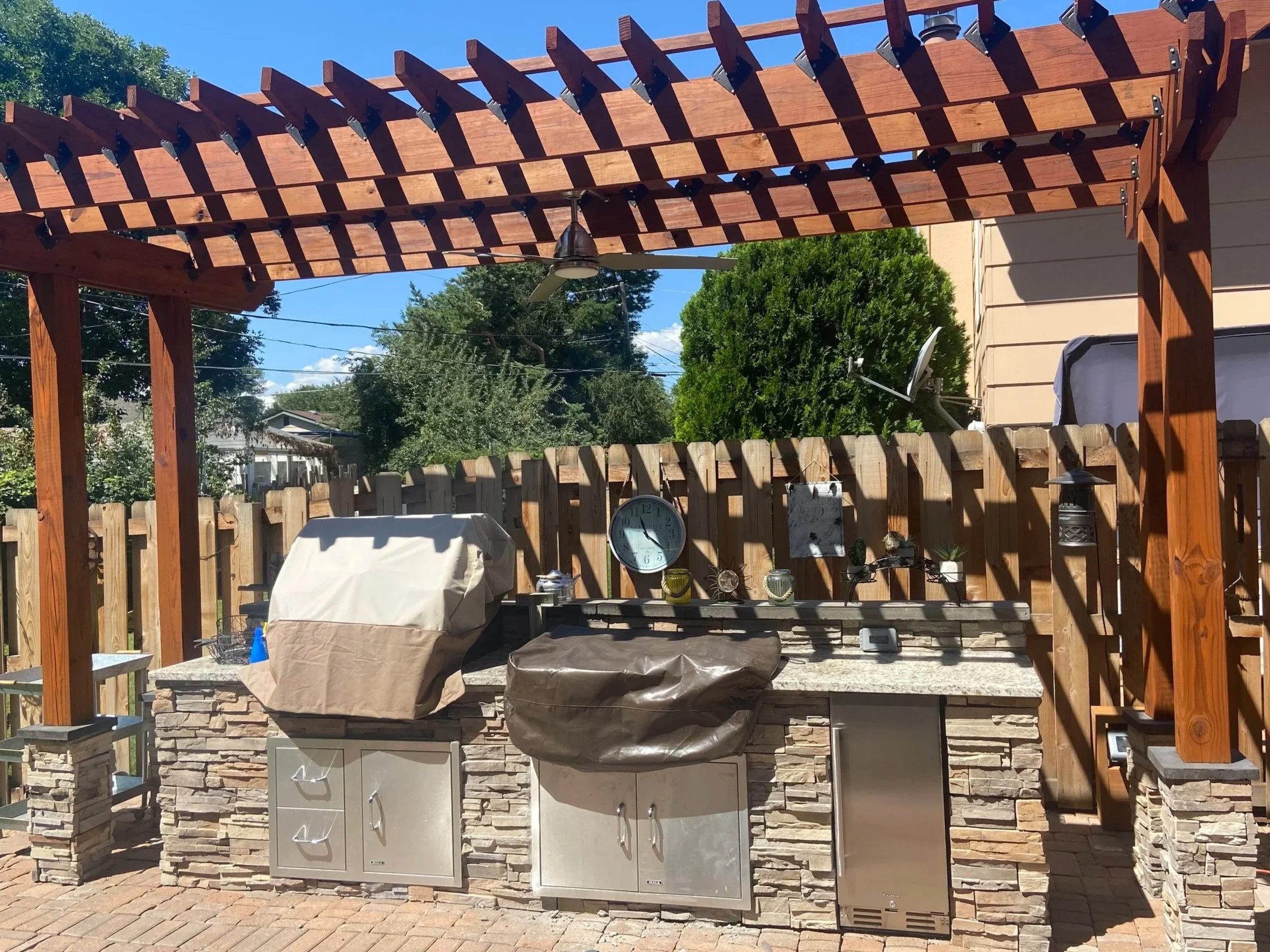 Wooden pergola over outdoor kitchen with gas grills, stone veneer, wall clock, satellite dish, and potted plants in backyard, under sunny blue sky.