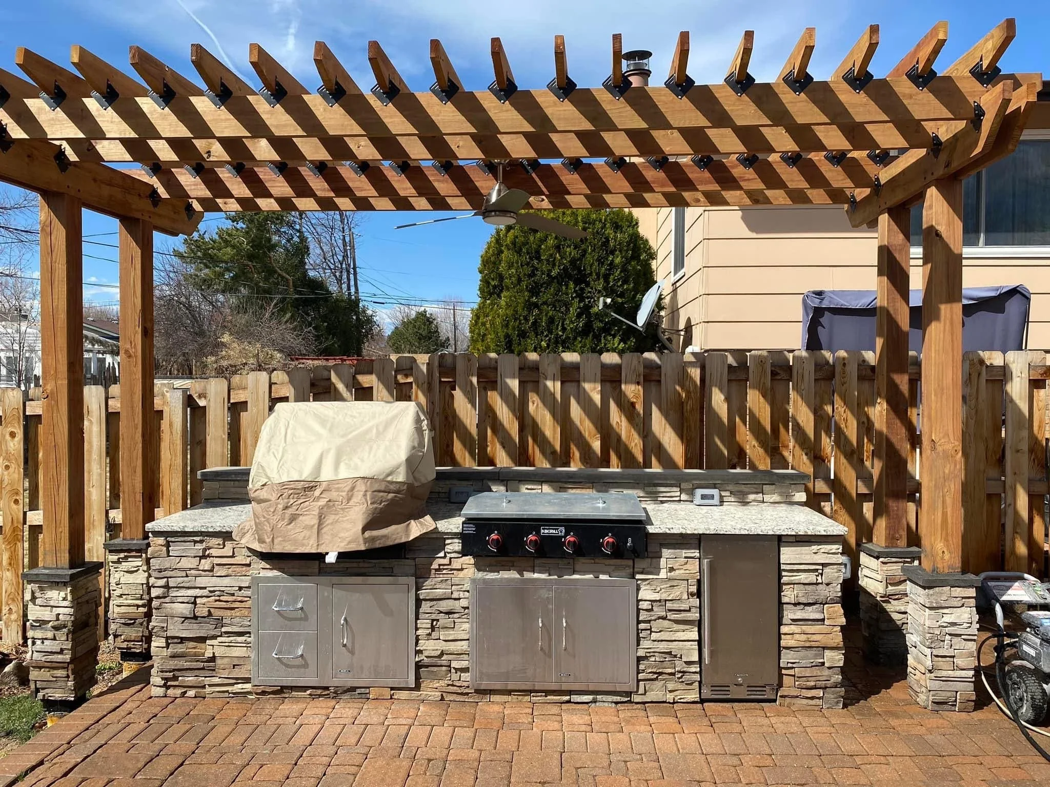 An outdoor kitchen area with a stone countertop, a covered grill, and a wooden pergola structure with a ceiling fan. There is a beige house with a siding exterior in the background and a wooden privacy fence surrounding the space.