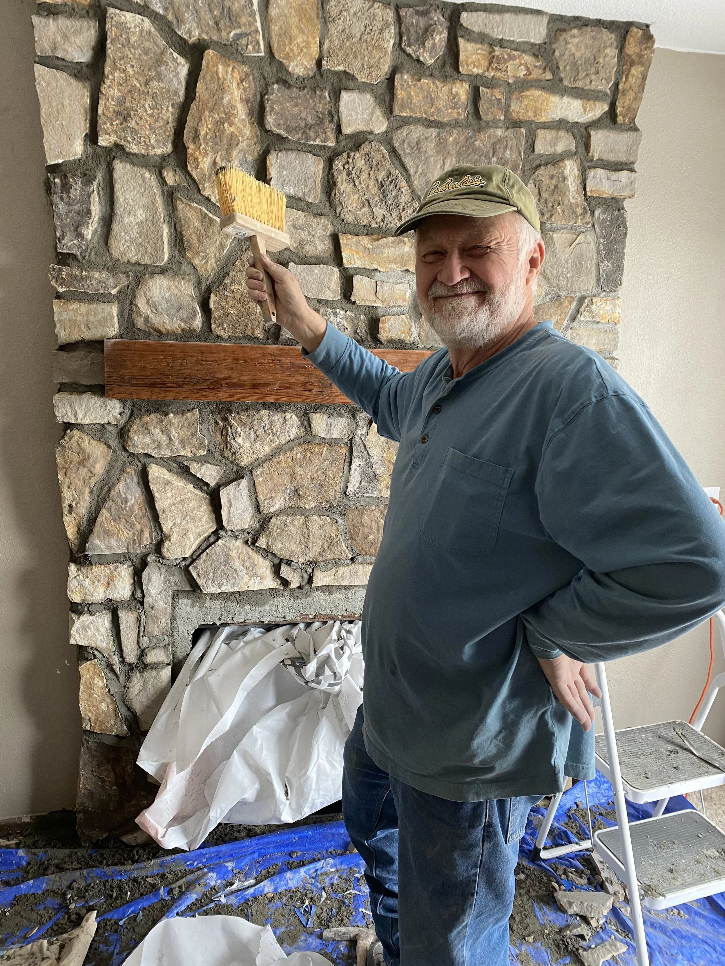 Older man smiling and holding a paintbrush in front of a partially finished stone and wood fireplace. He is wearing a green cap and a blue shirt, standing on a blue tarp with tools and materials around.