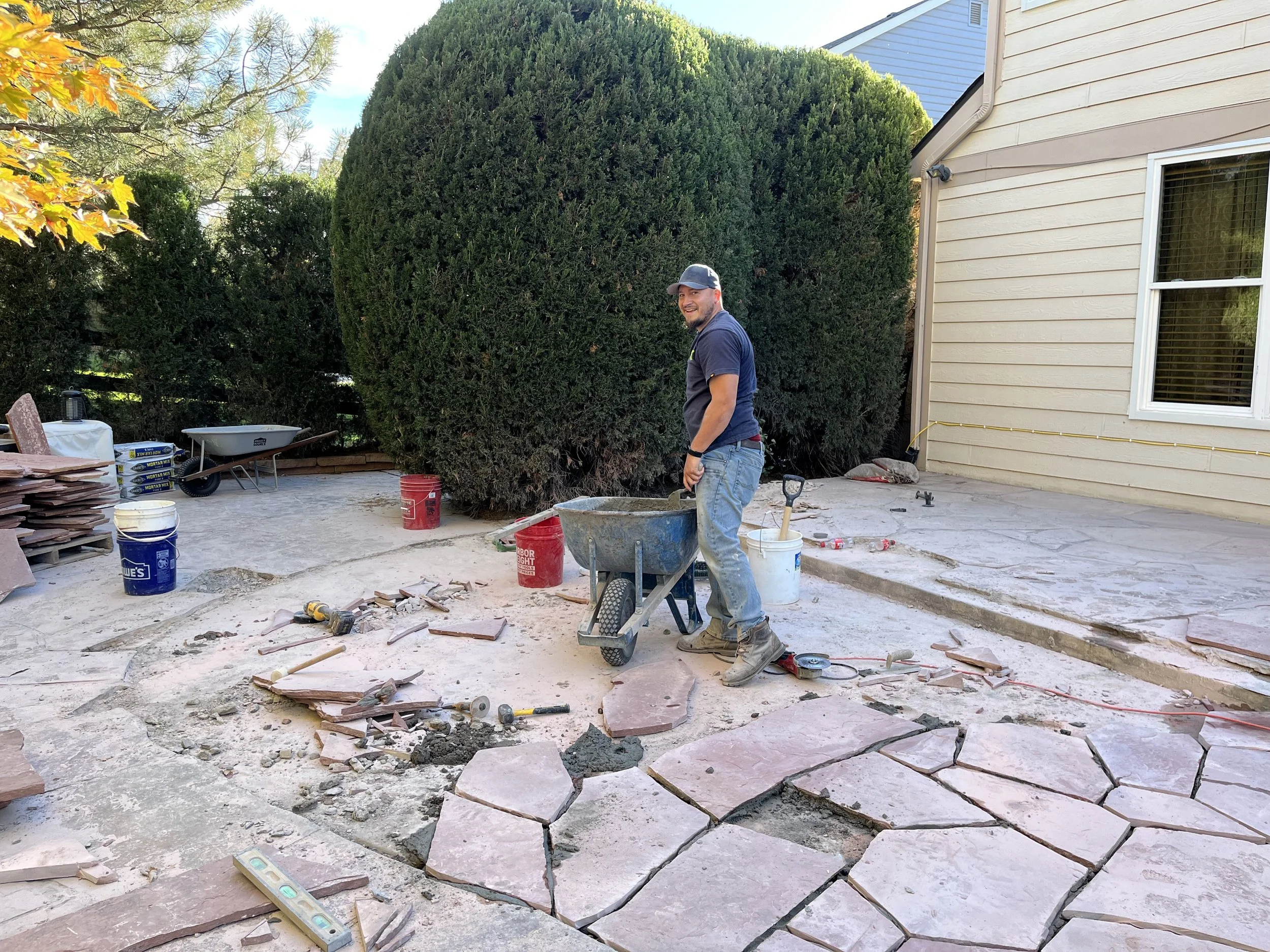 A man wearing a cap, gray t-shirt, and jeans working on a patio renovation, surrounded by construction materials, tools, and a partially removed concrete patio with large cracks. Tall trees and the exterior wall of a beige house are in the background.