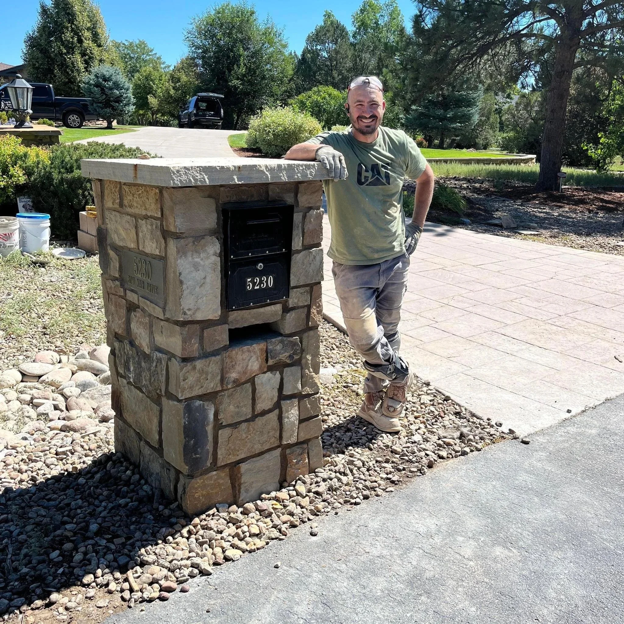 A man smiling and leaning on a stone mailbox in a residential yard on a sunny day.