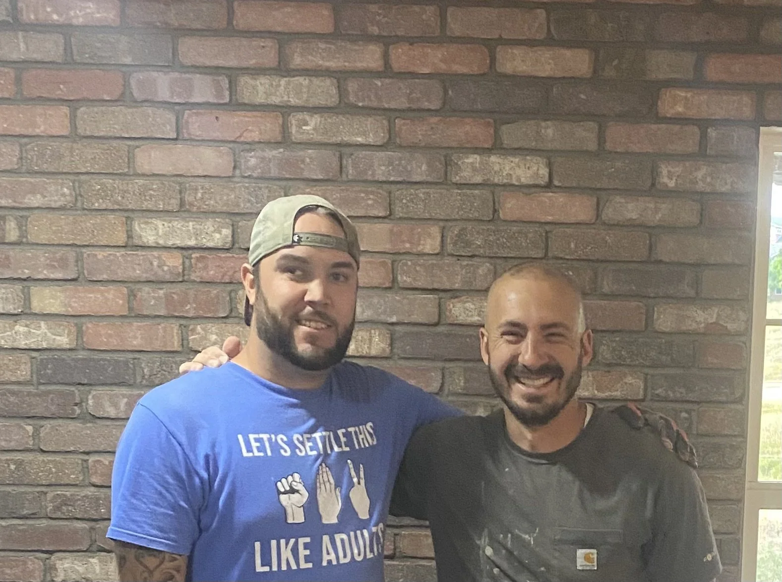 Two men standing in front of a brick wall, smiling and posing for the photo. One man wears a blue T-shirt with white text and a backwards baseball cap, the other wears a black shirt and has a buzz cut.