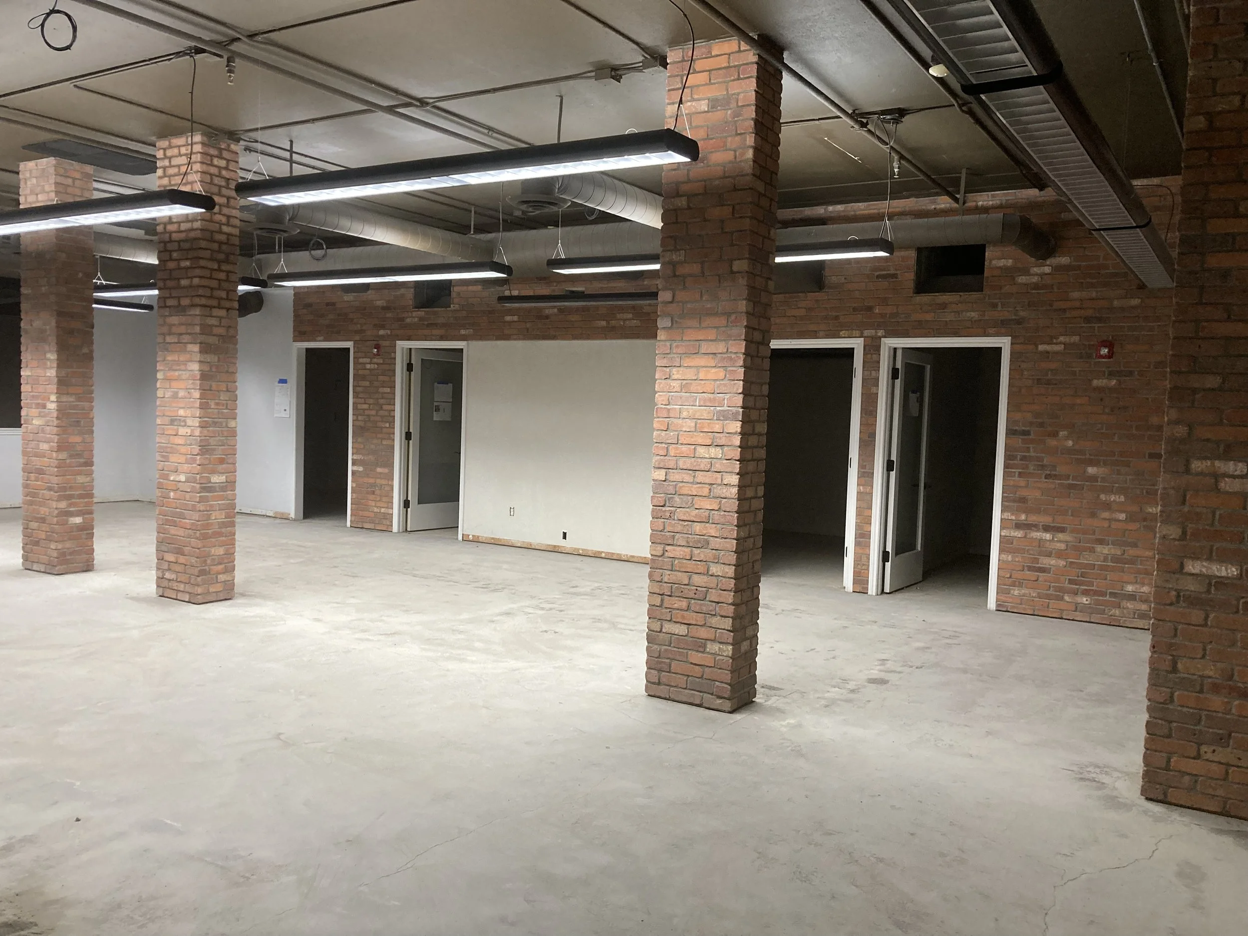 Interior of an unfinished commercial space with brick pillars, exposed ductwork, fluorescent lighting, and open doorways.