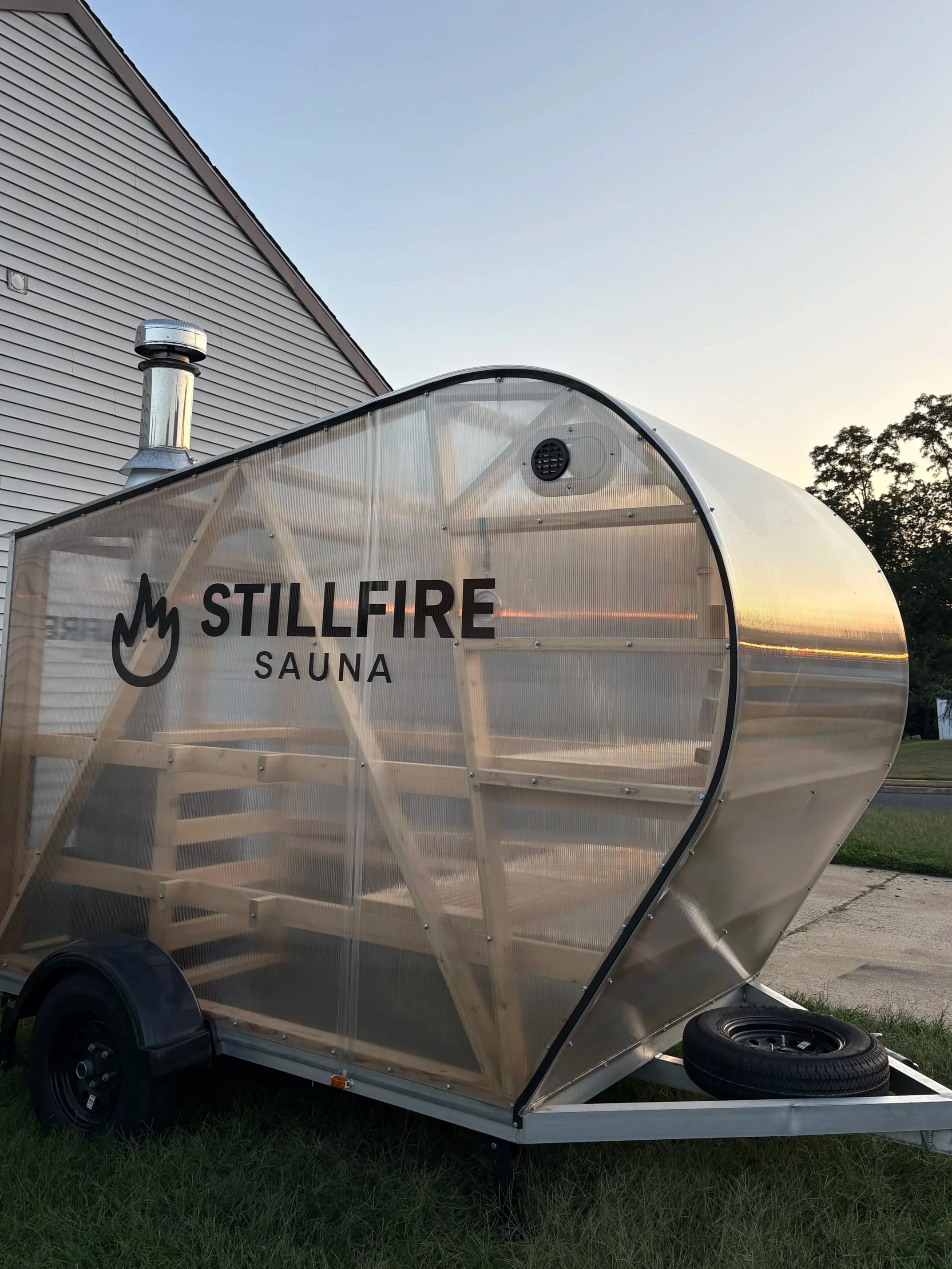 Image of a clear wood-fired mobile sauna parked in a backyard at sunset.