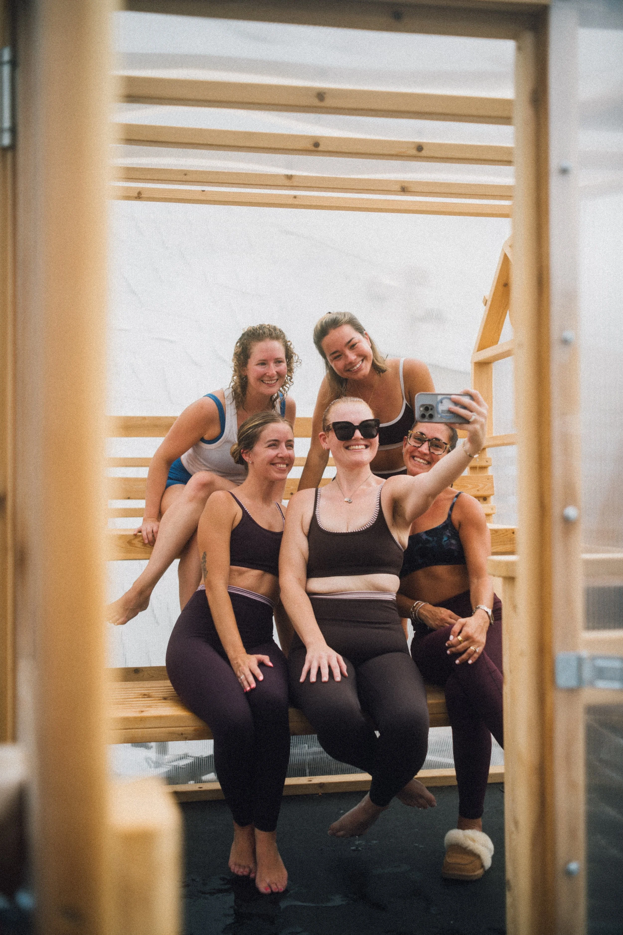 People sitting inside a small wooden modern looking sauna with a transparent door, during the day, taking a selfie on the wooden sauna benches. They appear to be smiling and enjoying the sauna.